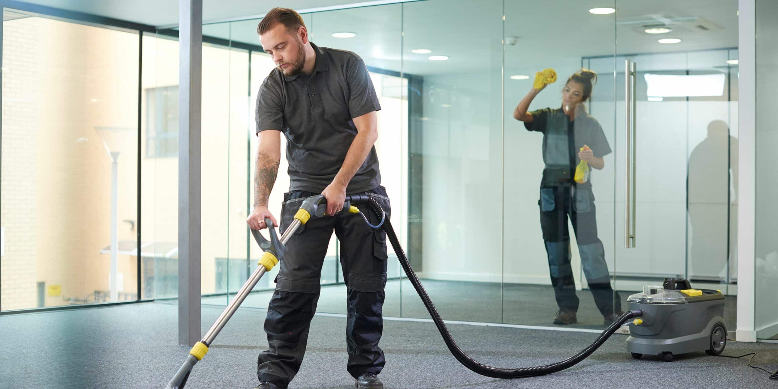 Man vacuuming a carpet floor and a woman cleaning a glass divider.