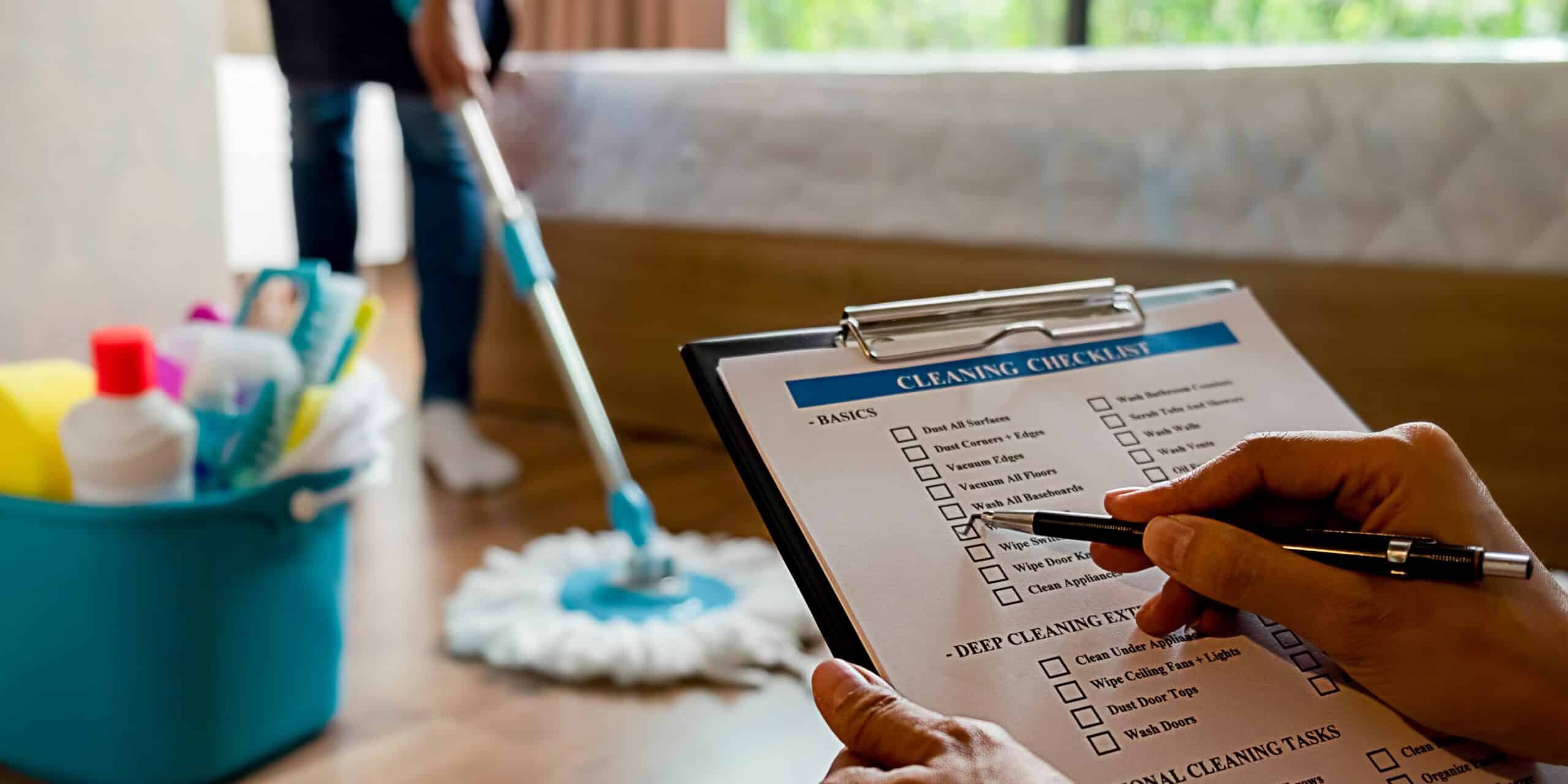 A hand holding a checklist of cleaning activities that needs to be done with a man mopping the floor on the background.
