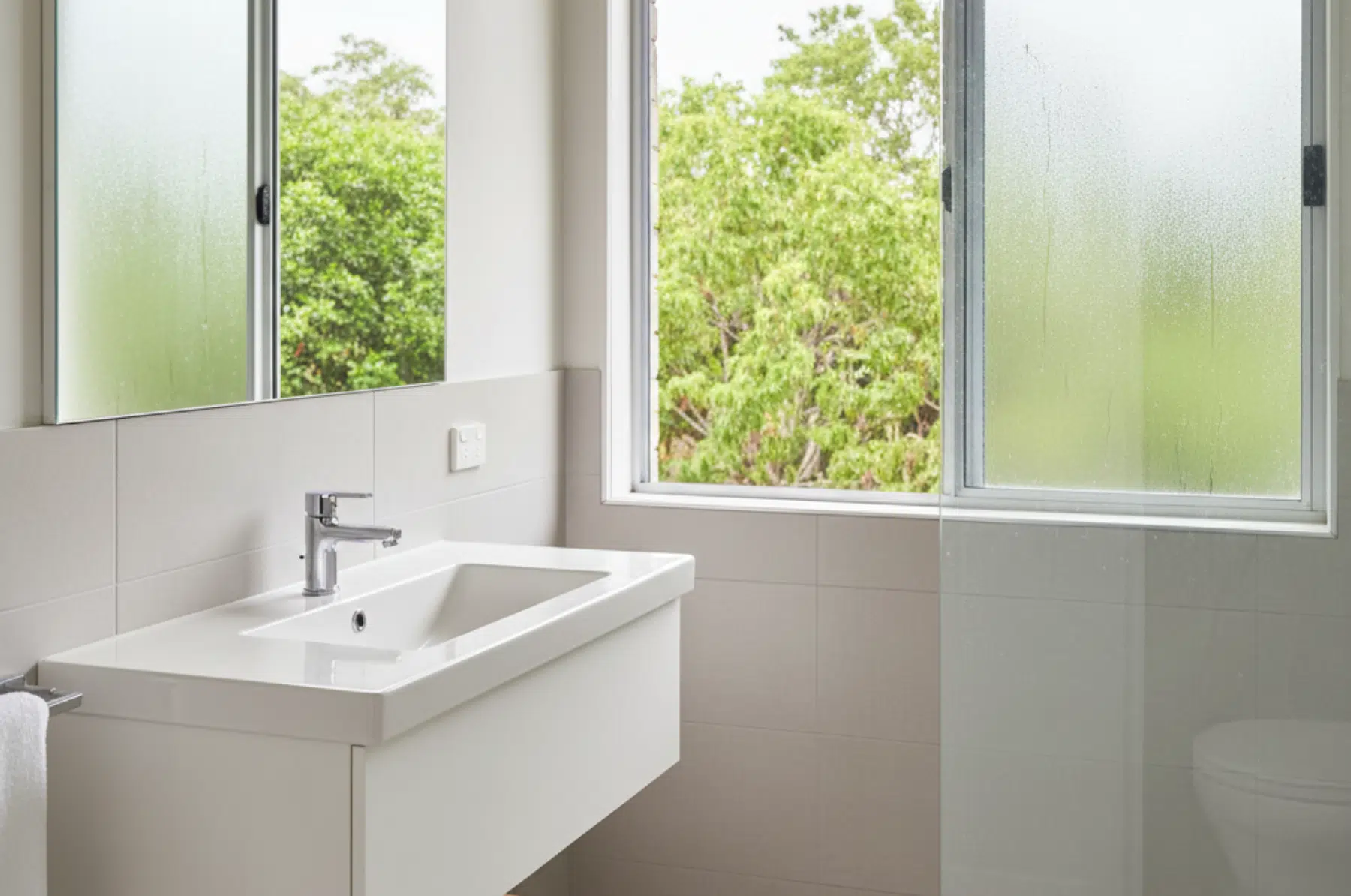Modern bathroom with a wall-mounted sink, large mirror, frosted window, and glass shower enclosure overlooking greenery.